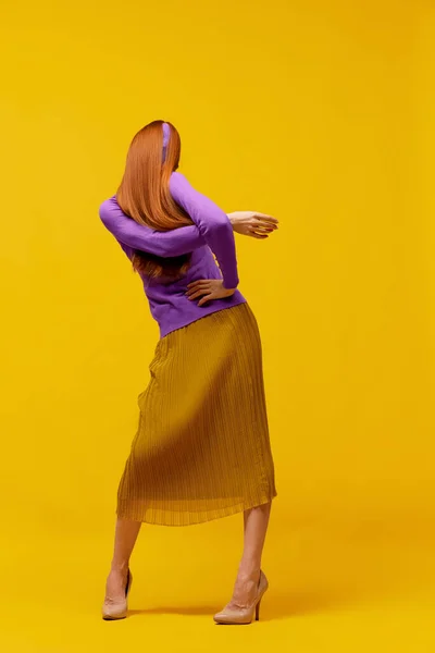 Weird dance. Faceless portrait of slim woman with long straight red hair wearing retro fashion outfit in action over yellow studio background. Impersonal emotions, body language