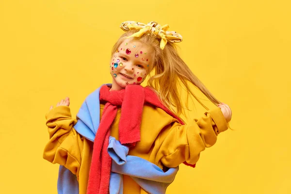 Enjoying, happiness. Portrait of little girl wearing bow on head touching blond hair and smiling to camera over yellow background. Concept of fashion, ad, childrens products, child model