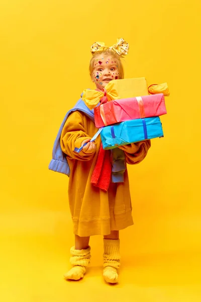 Surprised, gift, happy birthday. Full-length photo of little girl holding gifts, boxes with surprised face over yellow background. Concept of celebration, ad, childrens products, childhood, family