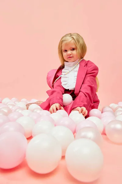 Lets play. Photo of little, stylish girl with blond hair wearing pink clothes and playing with balls over pink background. Concept of childhood, childrens games, child model, ad