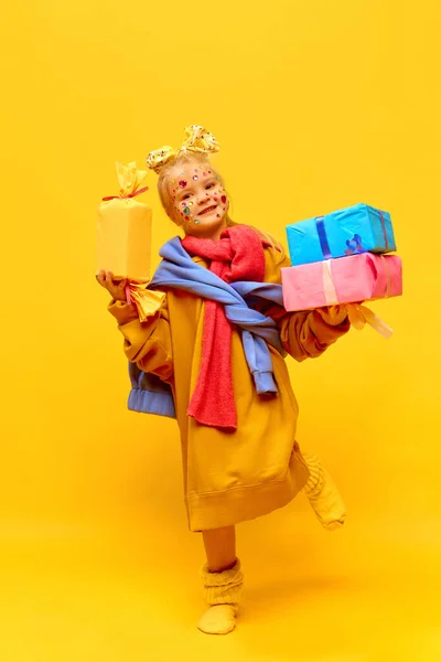 Happiness, gift, happy birthday. Full-length photo of little girl holding gifts, boxes with happy face over yellow background. Concept of celebration, ad, childrens products, childhood, family