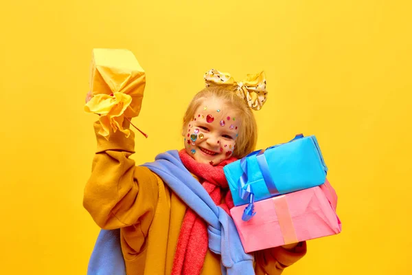 Happiness, gift, happy birthday. Portrait of cheerful little girl holding a lot of gifts, boxes with happy face over yellow background. Concept of celebration, ad, childrens products, childhood