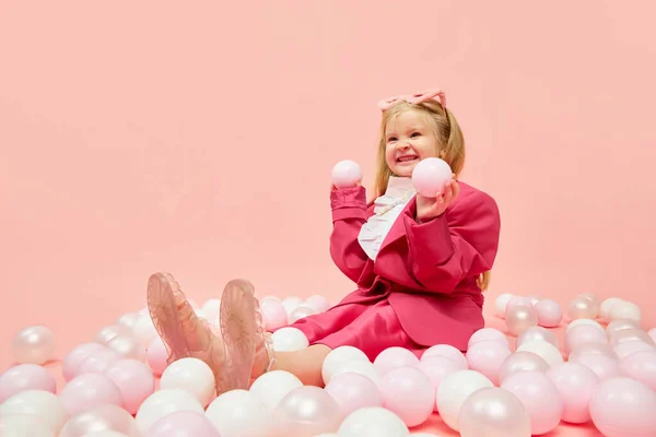 Fun, games. Photo of little girl with blond hair wearing pink clothes and sunglasses, playing with balls with hands over pink background. Concept of emotions, childhood, childrens games, ad