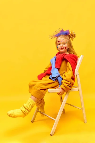 Surprised, happy emotions, happiness, fun. Photo of sweet little girl wearing bow, sitting on chair and laughing over yellow background. Concept of emotions, childhood, child model, ad