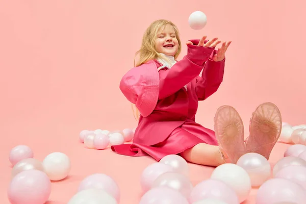 Happiness, fun. Photo of little child with blond hair wearing pink clothes playing, throwing ball and smiling over pink background. Concept of emotions, childhood, childrens games, ad