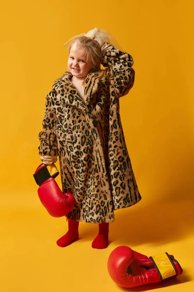 Photo of little blond stylish girl wearing boxing gloves and leopard print furcoat standing and looking away over yellow background. Concept of human emotions, gestures, fashion, ad, child model
