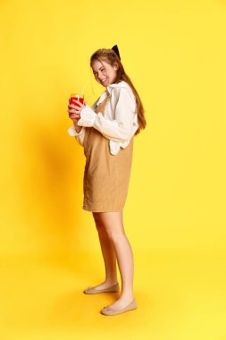 Neşeli, mutlu duygular. Beyaz bluz giyen, elinde içki bardağı tutan ve sarı arka planda gülümseyen genç kızıl saçlı kızın portresi. Pozitif duygular, öğrenci hayatı, moda kavramı