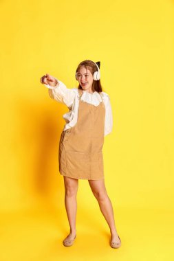 Neşeli duygular. Kulaklık takan, kamerayı işaret eden ve sarı arka planda dans eden duygusal genç ve güzel bir kız. Vay canına! Duygular, öğrenci hayatı, moda, müzik, reklam