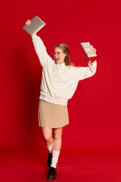 Success emotions. Excited student wearing costume, holding notebooks and keeping hands up over red background. Concept of student life, studying, learning, abroad educational