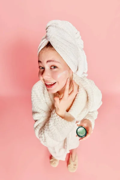 Morning beauty routine. Top view of cute beautiful girl wearing bathrobe shoulders using face cream to moisturize skin over pink background. Concept of beauty, natural make-up, skin care, healthy look