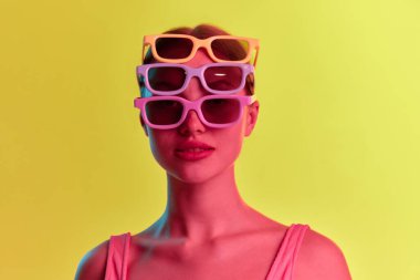 Moda. Birkaç güneş gözlüğü takan genç bir kızın portresi. Sarı-turuncu arkaplanda, neon ışıkta kameraya bakıyor. Güzellik kavramı, gençlik, insani duygular, ruh hali, reklam