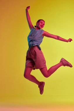 Özgürlüğü hissetmek. Zıplayan genç bir kızla el ele tutuşup neon ışıkta sarı-turuncu arka planda gülümseyen bir stüdyo fotoğrafı. Gençlik kavramı, mutluluk, ruh hali, boş zaman, reklam