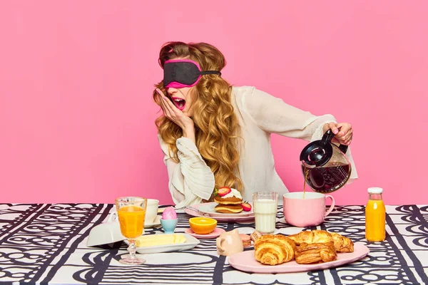 Lazy morning, french croissants, cooking meal. Sleepy beautiful girl with curly hair wearing sleep glasses preparing tea, breakfast and yawning over pink background. Concept of morning, food, ad
