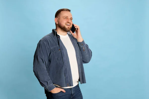 Smiling, communicative. Bearded man in jeans jacket and white t-short talking via cell phone, smiling and looking away over blue background. Concept of emotions, online work, sales, business
