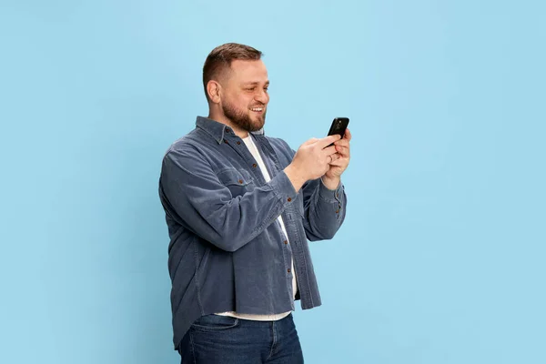 穿着牛仔裤夹克的快乐的留着胡子的男人看着手机 脸上挂着蓝色背景的笑容 情感概念 面部表情 在线工作 — 图库照片