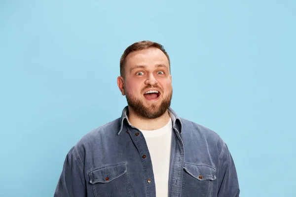 Surprise. Bearded handsome bearded man wearing jeans jacket smiling with surprised face and looking at camera over blue background. Concept of emotions, facial expression, mood