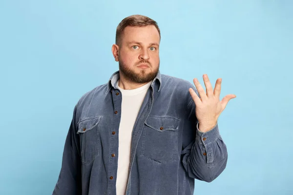 No idea, no clue. Good-looking man wearing jeans jacket spreading hand and looking at camera with face without understanding over blue background. Concept of emotions, facial expression, mood