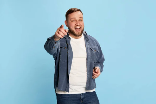 Choose lucky man. Lively handsome bearded man wearing white t-shirt pointing with finger at camera and smiling over blue background. Concept of bright emotions, facial expression, mood, ad