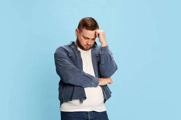 Problems, upset, life challenge. One pensive man standing with folded hands and thinking with sad facial expression over blue background. Concept of facial expression, mood, psychological condition