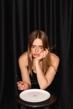 Toplum standartları. Tatminsiz genç kız, manken masada oturuyor ve neredeyse boş tabakları siyah arka planda yemeye başlıyor. Diyet, güzellik, mankenlik, yeme bozukluğu kavramı