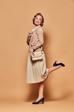 Sevinçli. Geçmişe dönük kıyafetler giyen, gülümseyen, bacağını eğen ve bej arka planda kameraya bakan güzel bir kadın. İnsani duygular, moda, güzellik, eski tarz, eski tarz, aktris