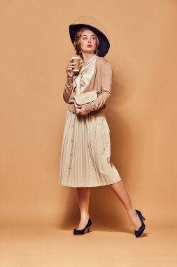 Kahve al. Retro giysiler giymiş güzel bir kadın elinde kahve fincanıyla bej arka plana bakıyor. İnsan duygularını kavra, güzellik, moda, retro stil, vintage, 60 'lar