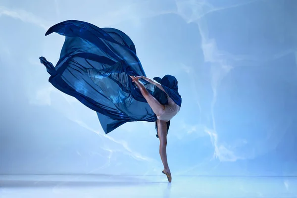 Tender soul. Young classical dancer dancing on fingertips with silk fabric over blue background. Concept of classic ballet, inspiration, beauty, dance, creativity. Beauty of contemporary dance