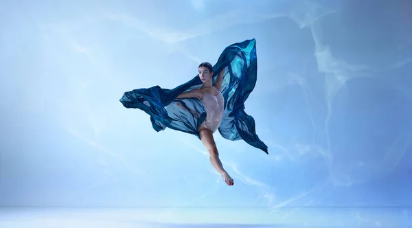 Flying with silk. Shot of jumping ballerina with stretched feet over blue background. Art, motion, action, flexibility, inspiration concept. Classic ballet. Beauty of contemporary dance