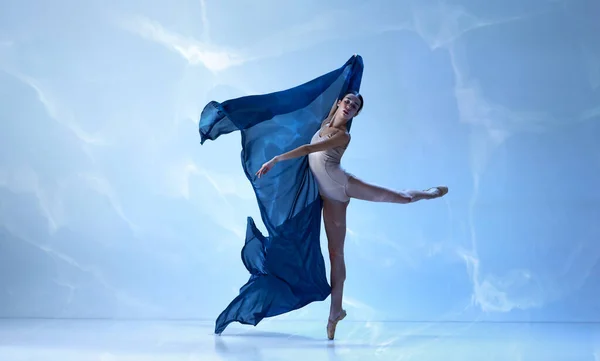 Solo performance. Ballerina dancing graceful movement with silk fabric over blue background. Art, motion, action, flexibility, inspiration concept. Beauty of contemporary dance