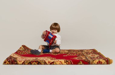 Eski tarz kıyafetler giyen mutlu çocuk halının üzerinde oturuyor ve armonikada hafif arka planda çalıyor. Dikkatsiz çocukluk kavramı, boş zaman aktiviteleri, eğlence, hobi, aile, eski tarz