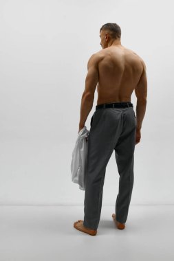 Klasik pantolon giyen ve beyaz arka planda üstsüz poz veren çekici bir adamın stüdyo fotoğrafı. Kaslı vücut şekli. Erkeklerin moda, tarz, vücut estetiği, güzellik, erkek sağlığı, duygu kavramı