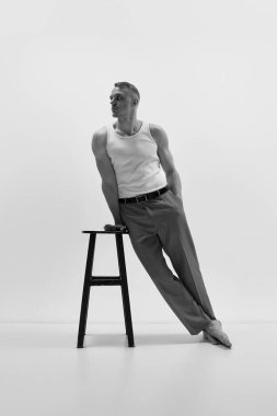 Pantolon giyen çekici kaslı adamın portresi ve stüdyo arka planında sandalyeye yaslanmış beyaz tişört. Siyah ve beyaz. Erkeklerin moda, tarz, vücut estetiği, güzellik, erkek sağlığı kavramı