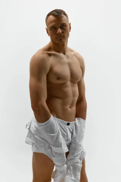 Aesthetics of male body. Portrait of handsome young muscular man posing shirtless over white studio background. Masculinity and strength. Concept of mens health, beauty of male body