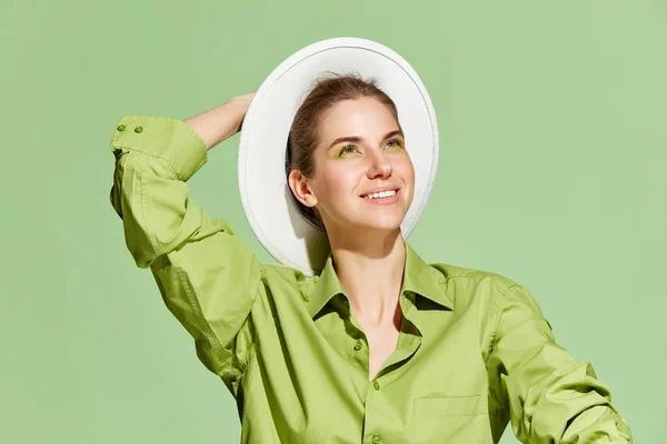 Summer, spring lifestyle clothes. Cheerful young adorable woman with bright makeup wearing hat and shirt, smiling over green background. Concept of fashion, style, beauty, wellness, skincare