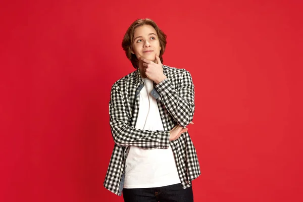 Let me think, thoughtful, doubtful. Half-length portrait of young boy in casual style clothes looking at camera with pensive face over red studio background. Concept of emotions, facial expression