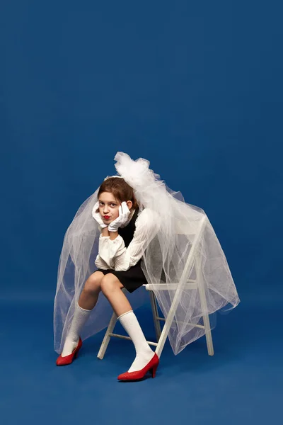 Little sad bride. Portrait of young girl wearing brides veil sitting on chair with sad facial expression over blue background. Concept of human emotions, mood, art, modern fashion, uniqueness