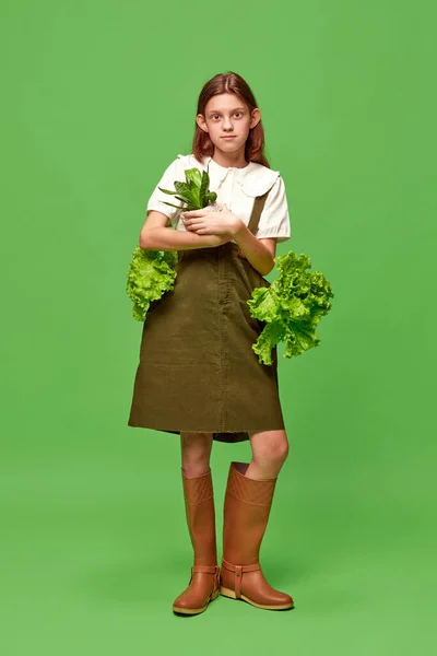 First harvest at garden. Shot of one adorable little girl in gardening clothes holding salad greens and smiling over green background. Concept of beauty, human emotions, mood, art, modern fashion, ad