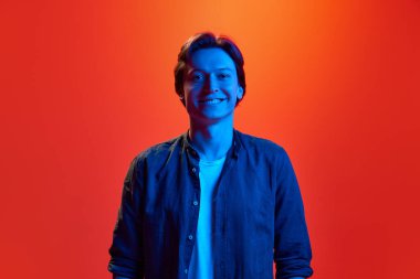 Geniş çapta pozitif. Kırmızı-turuncu arka planda neon ışıkta gülümseyen genç bir çocuğun stüdyo fotoğrafı. Güzellik kavramı, gençlik, insan duyguları, ruh hali, yüz ifadesi, reklam