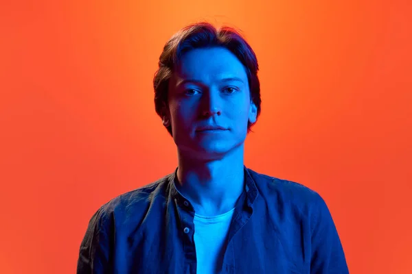Emotionless observer. Portrait of young man, guy with calm face looking away over red-orange background in neon light. Concept of beauty, youth, human emotions, mood, facial expression, ad