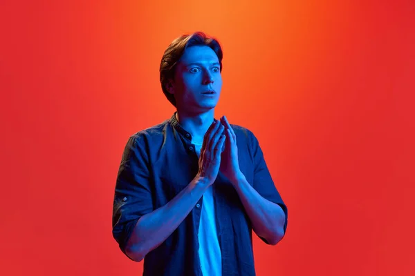 Hold your breath. Portrait of young funky guy, man wearing shirt standing with shocked face, fear on red-orange background in neon light. Concept of youth, human emotions, mood, facial expression, ad
