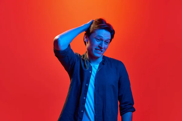 Doubtful guy. Portrait of young guy, man wearing shirt standing with uncertain facial expression over red-orange background in neon light. Concept of youth, human emotions, mood, facial expression, ad