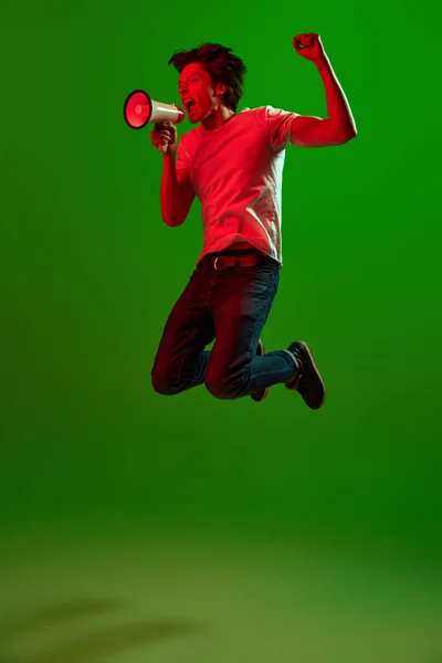 Breaking news. One young angry man, guy screaming through megaphone, jumping and threatens with a fist over green background in neon light. Concept of information, social media, human emotions, ad
