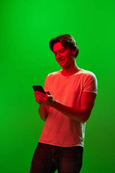 社交网络 在线聊天 穿着白色T恤的快乐年轻人看着手机 对着绿色霓虹灯的背景笑着 情感概念 面部表情 在线工作 — 图库照片