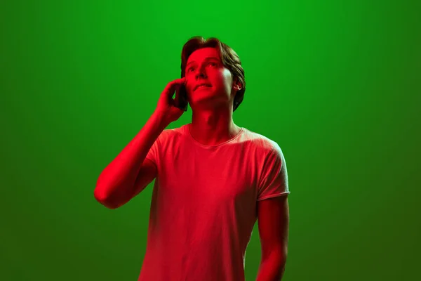 Long distance relationship. Attractive man wearing white t-shirt talking via cell phone and looking away over green neon background. Concept of emotions, online work, sales, business