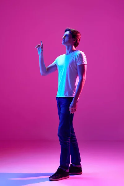 白いTシャツを着て ネオンの光の中でピンクの背景に指を指している若いハンサムな男 サイドビュー ゲーム デジタル化 テクノロジーの概念 内なる世界 — ストック写真
