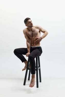 Kas şeklindeki vücut şekli. Beyaz stüdyo arka planında üstsüz poz veren çekici genç bir adam. Moda, tarz, vücut estetiği, güzellik, erkek sağlığı kavramı. Model sakin ve sakin görünüyor.