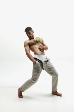 Romantik hareket. Beyaz stüdyo arka planında çiçeklerle üstsüz poz veren çekici genç bir adam. Moda, tarz, vücut estetiği, güzellik, erkek sağlığı kavramı. Model sakin ve sakin görünüyor.