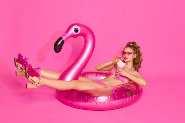 stock image Pool party. Portrait of blond girl wearing swimsuit drinking milkshake and laying on inflatable flamingo over pink studio background. Concept of summer, vacation, party, fashion, beauty, youth, ad