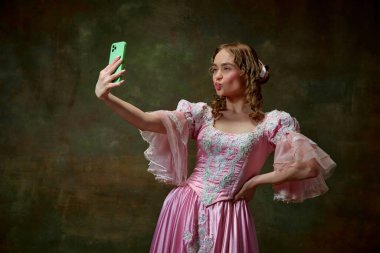 Ördek dudaklı selfie. Güzel sarışın prensesin portresi, süslü pembe elbiseler giyerek ve tablet üzerinde özçekim yaparak klasik doku arka planında. Ortaçağ, güzellik, eski moda kıyafetler.