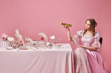Tanrıların yiyeceği. Peri masalı sarışın kraliçe, Rönesans elbisesi giyen prenses pembe stüdyo arka planındaki şenlik masasının yanında büyük bir sandviç tutuyor. Ortaçağ, güzellik, yemek, barok, reklam kavramı.